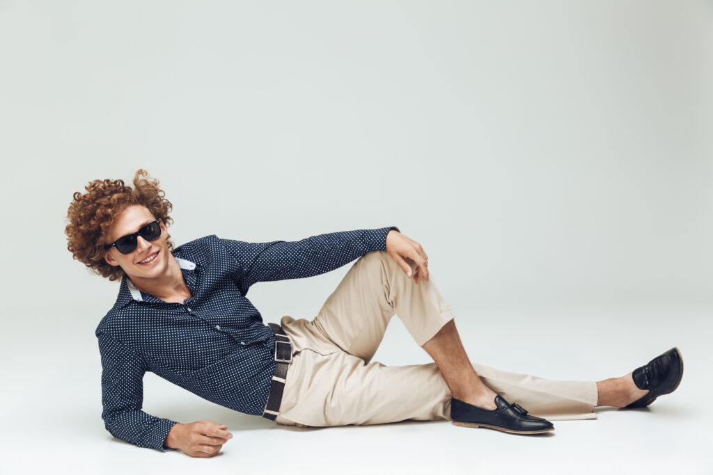 Man with curly hair wearing sunglasses, a navy patterned shirt, and beige trousers, reclining casually on the floor against a plain background. Presenting the workwear capsule wardrobe.